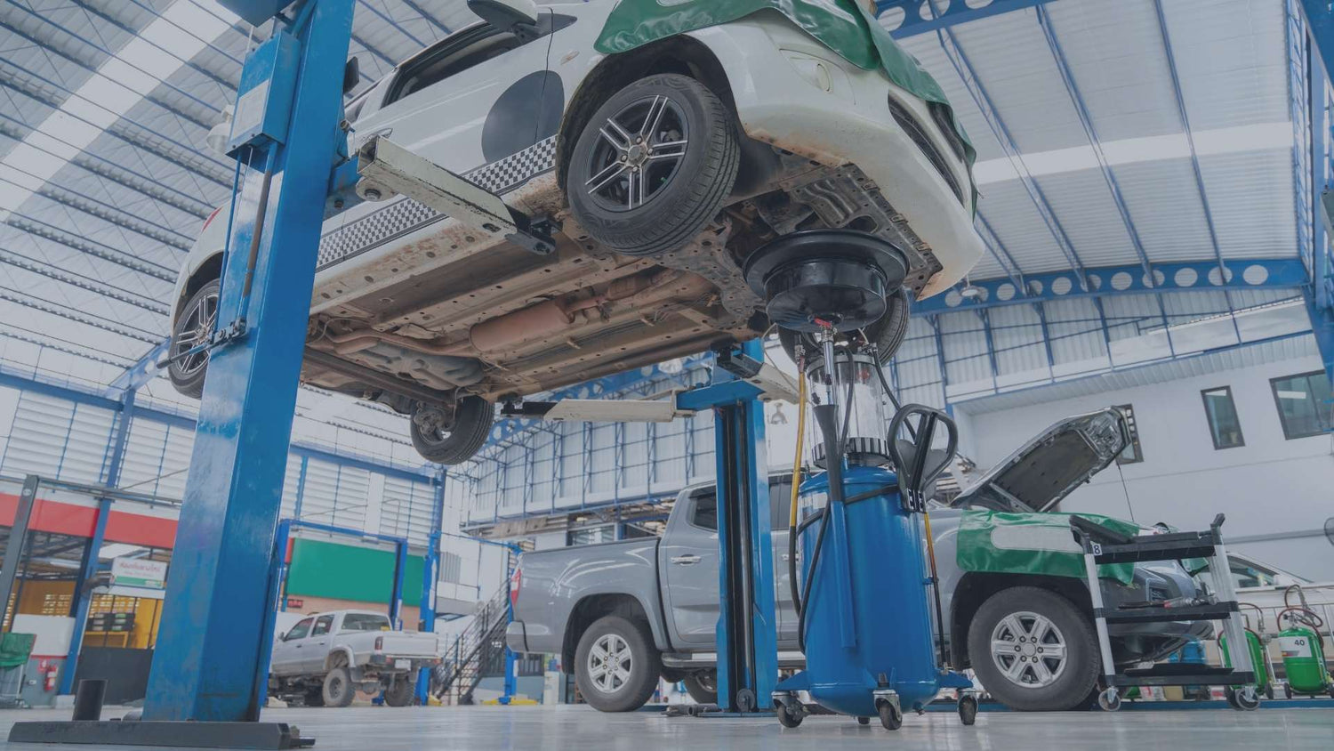 Blick in eine moderne Kfz-Werkstatt mit einer professionellen blauen Zweisäulen-Hebebühne, auf der ein weißes Auto für Wartungsarbeiten hochgefahren ist, daneben ein mobiles Altöl-Auffanggerät.