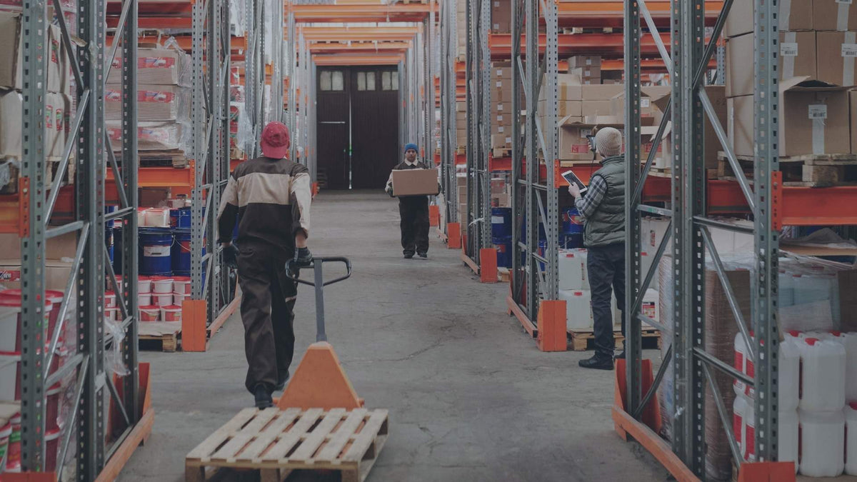 Drei Lagerarbeiter bei der Arbeit in einem gut gefüllten Logistikzentrum. Im Vordergrund zieht ein Mitarbeiter einen leeren Hubwagen, während ein Kollege rechts den Warenbestand mit einem Tablet und einem Scanner-Gerät kontrolliert.