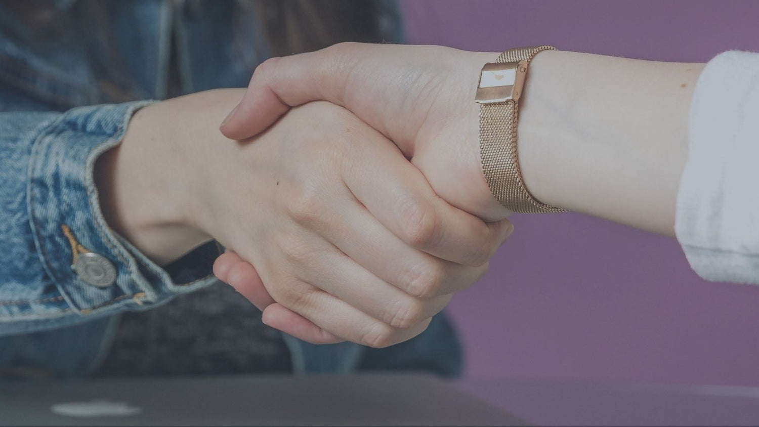 Eine Nahaufnahme von zwei sich schüttelnden Händen vor einem lilafarbenen Hintergrund. Die linke Person trägt eine Jeansjacke, die rechte Person trägt ein weißes Oberteil und eine goldene Armbanduhr am Handgelenk.