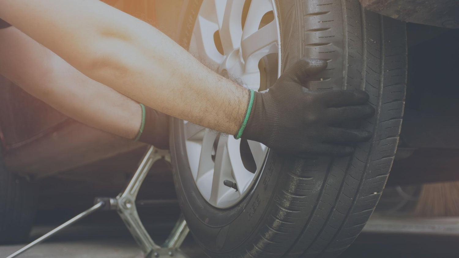 Nahaufnahme einer Person mit schwarzen Arbeitshandschuhen, die mit beiden Händen einen Reifen an einem Auto greift, das von einem klassischen Scherenwagenheber gestützt wird.