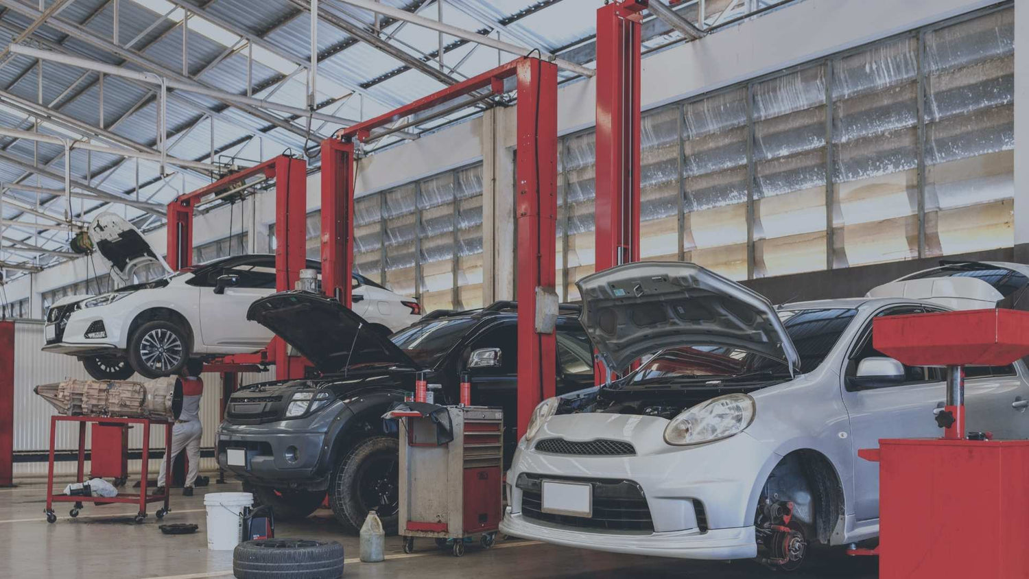 Eine große, helle Kfz-Werkstatt im laufenden Betrieb. Mehrere Autos stehen zur Reparatur an roten Hebebühnen bereit, teils mit geöffneter Motorhaube, während ein Mechaniker im Hintergrund unter einem angehobenen weißen Fahrzeug arbeitet.
