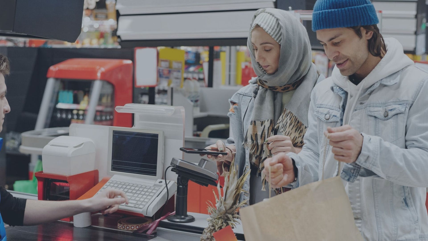 Eine Kundin bezahlt kontaktlos mit ihrem Smartphone an einem modernen POS-Kartenterminal in einem Supermarkt, während im Hintergrund das Kassensystem für den Bezahlvorgang bereitsteht.