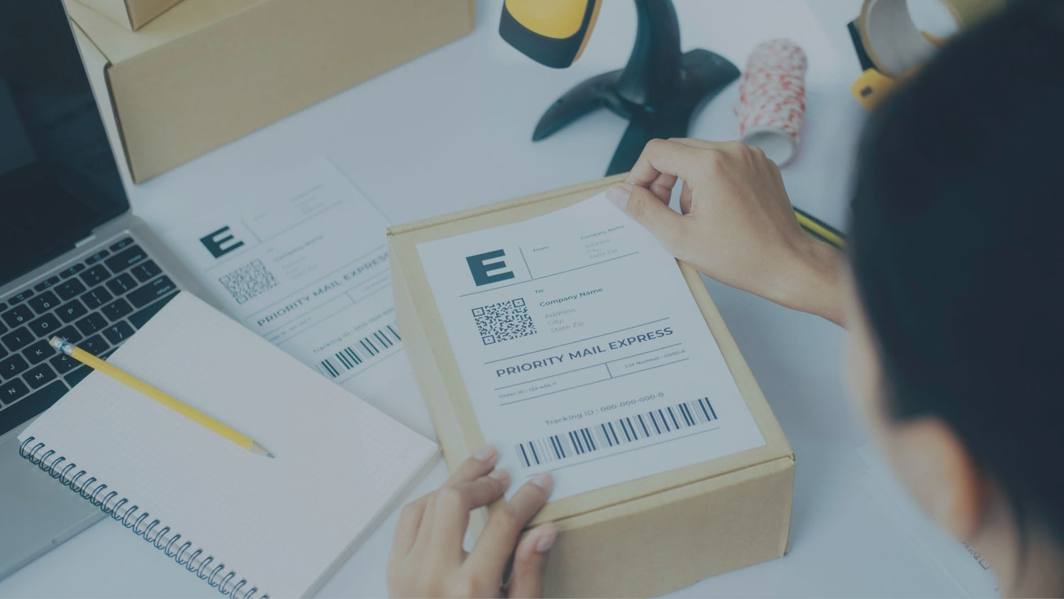 Blick über die Schulter einer Person an einem Packtisch, die ein ausgedrucktes Versandetikett mit Barcode auf einen kleinen Karton klebt. Im Hintergrund stehen ein Laptop, ein Notizblock und ein Barcodescanner in einer Standhalterung.